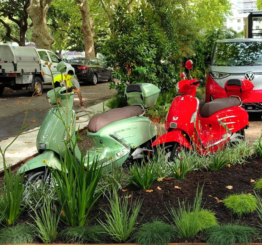 motorbikes, one green and one red, parked on a sidewalk with cars and trees in the background. at the Melbourne international flower show with Jamie durie